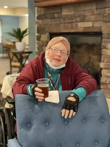 A woman in a headscarf and gloves sits in a wheelchair, holding a drink, in front of a stone fireplace—a warm scene reflecting women empowerment and strength in everyday life.
