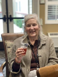 An older woman with gray hair sits indoors, smiling and holding a plastic cup with a brown beverage. She wears a brown shirt and patterned jacket, embodying the spirit of International Women’s Day and celebrating gender equality.