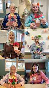Six older women sit and hold colorful handmade paper flower crafts with "I love you bunches!" tags, smiling at the camera in a decorated indoor setting, celebrating International Women’s Day and women empowerment together.