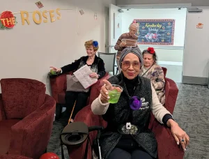 Four elderly adults sit in a lounge area with drinks at a festive Kentucky Derby watch party. One woman in the foreground raises her glass and smiles at the camera.