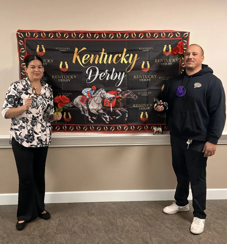Two people stand in front of a Kentucky Derby banner at a lively watch party, each holding a small trophy. The banner features horse racing imagery and decorative red flowers.