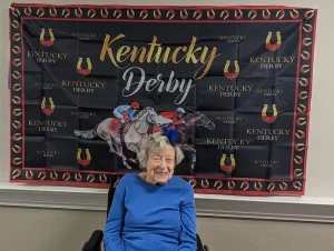 An elderly woman in a blue sweater sits smiling in front of a Kentucky Derby banner, ready to enjoy the watch party surrounded by images of racing horses and jockeys.