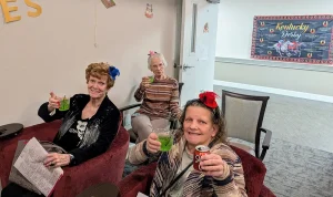 Three women seated indoors wear decorative hats, holding drinks and smiling at a lively Kentucky Derby Party. A Kentucky Derby poster is visible on the wall in the background, adding to the festive watch party atmosphere.