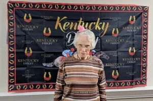 Elderly woman wearing a small pink hat stands in front of a Kentucky Derby Watch Party backdrop decorated with horse racing imagery and logos.