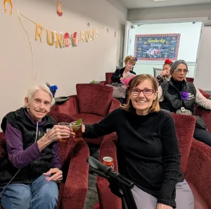 Five women sit in a lounge area with red chairs, two in the front raising drinks at a lively Kentucky Derby Party, with “Run for the Roses” decorations and a Kentucky Derby sign visible in the background.