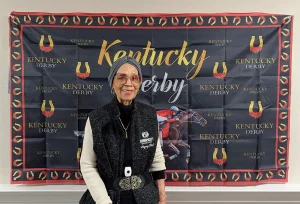 A woman stands in front of a Kentucky Derby Party banner featuring horses, horseshoes, and the event logo. She is wearing glasses, a gray headscarf, and a black-and-gray outfit at the festive watch party.
