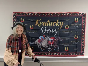 An older woman with a walker stands smiling in front of a Kentucky Derby banner featuring horses and jockeys at a lively Kentucky Derby Party.