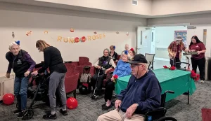 A group of elderly people, some with walkers and wheelchairs, gather in a decorated room with a "Run for the Roses" banner and a green table with red flowers for a festive Kentucky Derby Party.