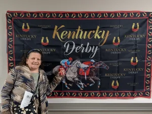 A woman stands in front of a Kentucky Derby backdrop featuring racehorses and jockeys, holding a can of soda and a piece of paper at a lively Kentucky Derby Party.