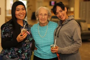 Three women stand together indoors, smiling at the camera. Two women hold cups with spoons, and the woman in the middle wears glasses and a blue top with a pearl necklace.