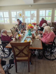 Several elderly women in wheelchairs sit around a decorated table in a bright room, enjoying community activities with flowers and gift bags on the table.