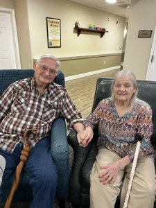 An elderly man and woman sit side by side holding hands on armchairs in a well-lit room, each with a cane, reflecting a warm sense of community and shared lifestyle.