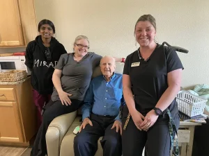 Three women and an elderly man are posing and smiling in a cozy room, reflecting a warm sense of community, with a couch, kitchen counter, and basket visible in the background.
