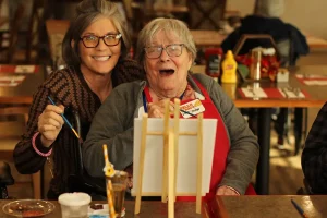 Two women sit together at a table with painting supplies, sharing smiles and creativity. Their joyful moment captures a lifestyle of artistic expression and community, with an easel, art materials, and condiments in the background.
