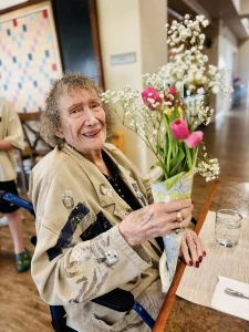 An elderly woman sitting in a wheelchair holds a bouquet of flowers and smiles, enjoying a warm community atmosphere at a table with a glass of water and a placemat, reflecting her joyful lifestyle.
