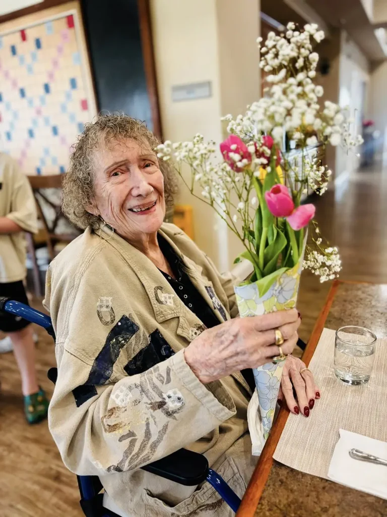 An elderly woman sitting in a wheelchair holds a bouquet of flowers and smiles, enjoying a warm community atmosphere at a table with a glass of water and a placemat, reflecting her joyful lifestyle.