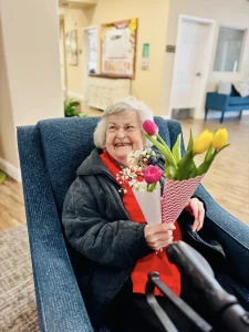 An elderly woman in a black jacket and red shirt sits in a blue chair, smiling and holding a bouquet of tulips and baby's breath flowers, capturing a warm moment of community and lifestyle.