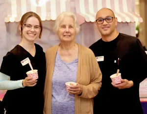Three people, two in black scrubs and one in casual clothes, stand together indoors, each holding a cup of ice cream and smiling at the camera.