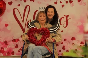 Two women celebrate Valentine's Day at a community event—one seated and holding a red heart decoration, the other standing behind her in a warm hug. The festive backdrop with hearts highlights the joyful lifestyle they share together.