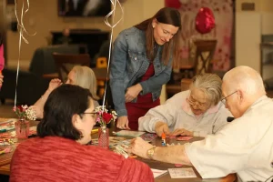 A group of older adults sit at a table doing crafts, embraced by a vibrant sense of community, as a younger woman assists them. Balloons and flowers decorate the table, creating a joyful lifestyle setting.
