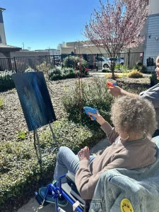 Embracing a vibrant lifestyle, two people—one in a wheelchair—work together as a community, using water guns to spray paint onto a canvas set on an easel outdoors on a sunny day.