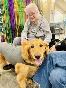 An elderly woman in a wheelchair smiles while petting a golden retriever wearing a gray vest, enjoying a warm moment of community. Bright streamers hang in the background, reflecting the joyful lifestyle of the gathering.