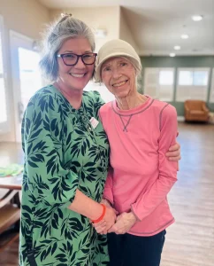Two women stand indoors, smiling and embracing each other—a snapshot celebrating community and lifestyle. One wears a green patterned dress and glasses; the other sports a pink shirt and a light-colored hat.