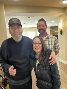 Three adults pose indoors, reflecting a sense of community and lifestyle—one man in a black cap and sweatshirt, another in a plaid shirt, and a woman in glasses and a dark jacket—all standing close together and smiling.