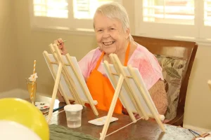 An elderly woman wearing an orange apron sits at a table and smiles while painting on a canvas, enjoying her creative lifestyle with art supplies, a drink nearby, and the warmth of her community.