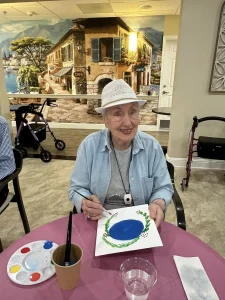 An elderly woman wearing a white hat sits at a table, painting on a canvas with blue paint, embracing a creative lifestyle in a room adorned with a European street mural and walking aids, reflecting her vibrant community spirit.