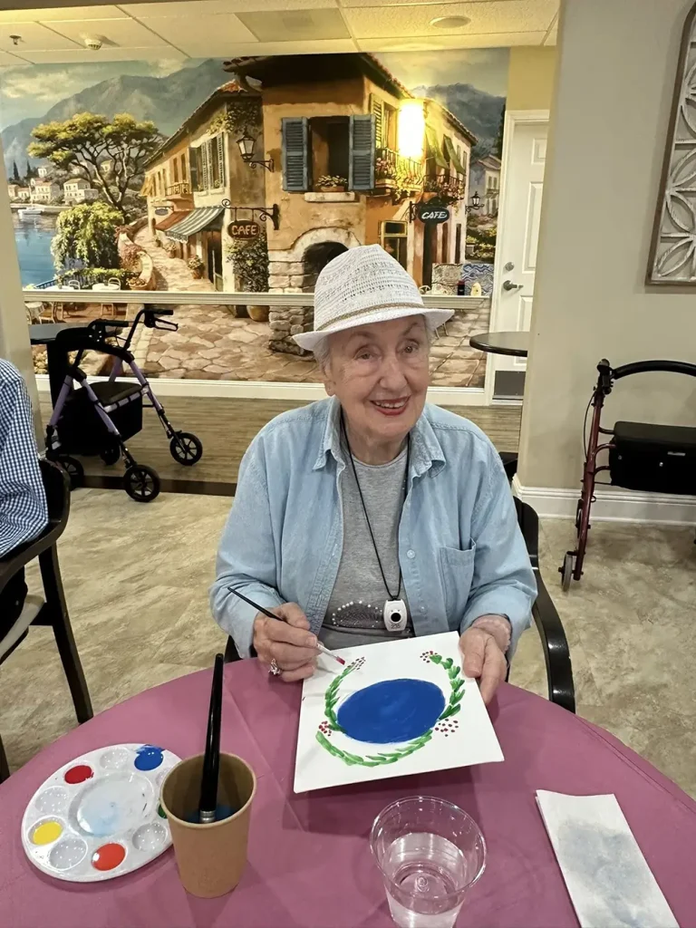 An elderly woman wearing a white hat sits at a table, painting on a canvas with blue paint, embracing a creative lifestyle in a room adorned with a European street mural and walking aids, reflecting her vibrant community spirit.