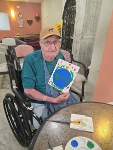 An elderly man in a wheelchair holds up a hand-painted picture of the Earth with colorful hearts, celebrating community and lifestyle as he sits at a table with painting supplies in a cozy room.