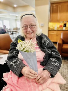 Elderly woman with glasses smiles while sitting indoors, holding a small bouquet of flowers wrapped in paper. She wears a dark cardigan with butterfly patterns and a pink skirt, radiating warmth and celebrating community lifestyle.