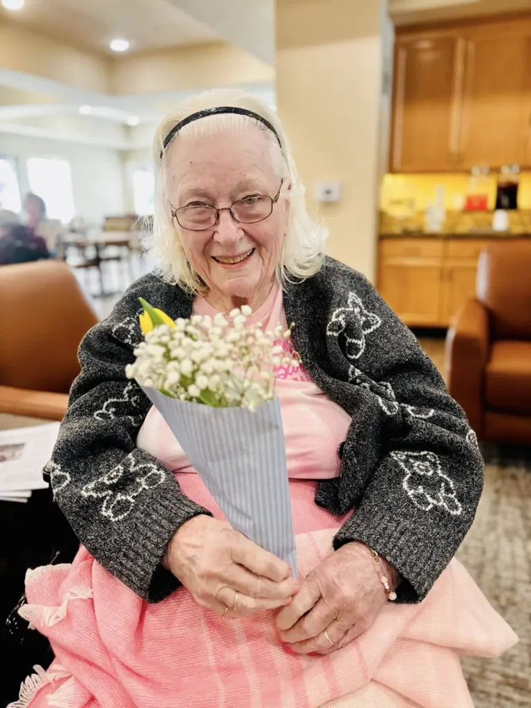 Elderly woman with glasses smiles while sitting indoors, holding a small bouquet of flowers wrapped in paper. She wears a dark cardigan with butterfly patterns and a pink skirt, radiating warmth and celebrating community lifestyle.