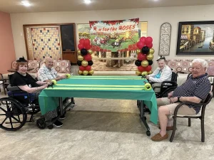 Four elderly adults sit around a table decorated with green cloth and balloons, enjoying a "Race for the Roses" themed indoor activity that celebrates community and an active lifestyle.
