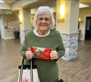 Older woman with gray hair and glasses, wearing a green shirt, holds a package of peanut butter cookies and a white tote bag, standing indoors with soft lighting—a warm moment reflecting community and lifestyle.