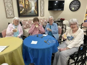 Four elderly women sit around a blue table, smiling and holding dessert cups, enjoying community and conversation in a cozy, well-lit room adorned with wall art, a TV, and a large clock in the background.