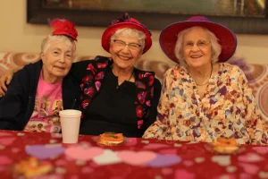 Three elderly women wearing colorful hats and smiling sit together at a table decorated with a heart-patterned tablecloth and plates of food, celebrating the joy of community and a vibrant lifestyle.