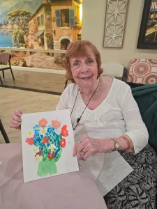 An older woman sits at a table, smiling and holding up a colorful painting of flowers in a vase, celebrating her creative lifestyle within the community.
