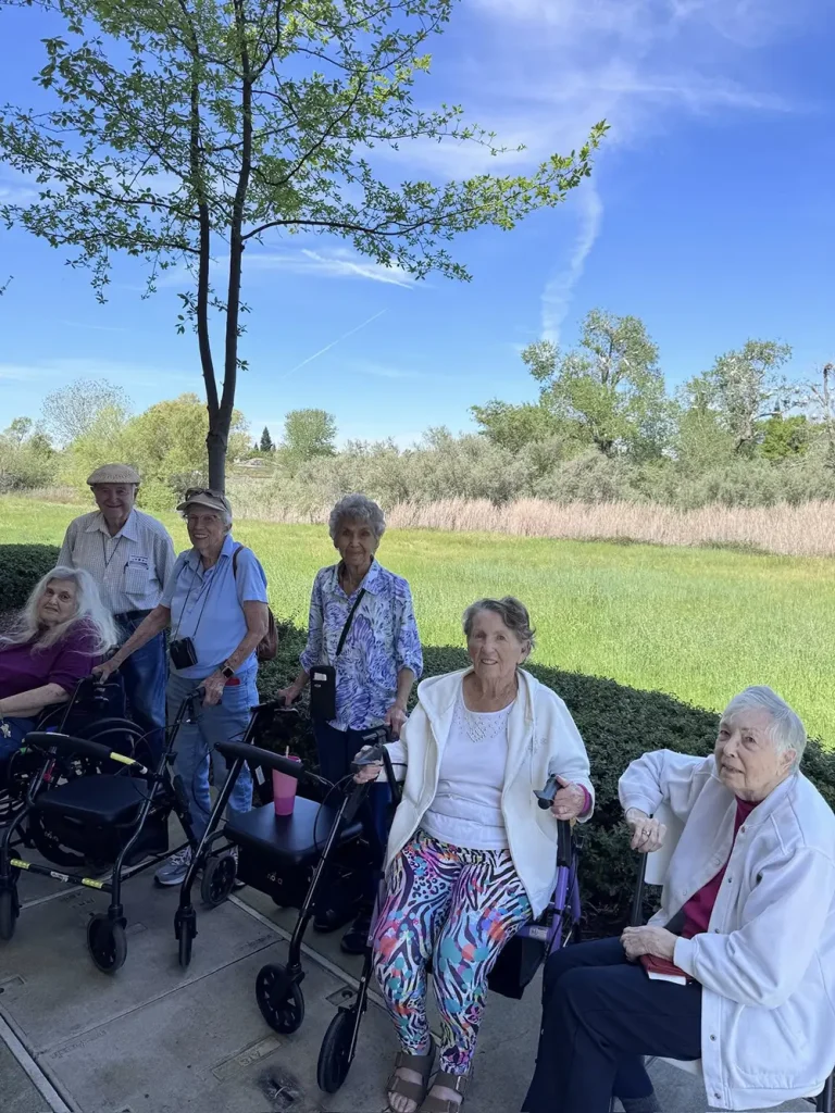 Six elderly people, some using walkers, are enjoying a vibrant community lifestyle as they sit and stand outdoors on a paved path near grass and trees under a blue sky.