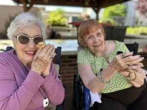 Two elderly women sit outside under a gazebo, each smiling and holding a small chick in their hands, enjoying a peaceful moment of community and lifestyle together.