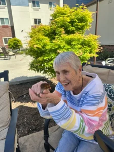 An elderly woman with short gray hair sits outdoors, smiling and gently holding a small bird in her hands, enjoying a peaceful lifestyle amid a vibrant community, with a green tree and buildings in the background.