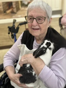 Elderly woman with glasses sitting and holding a black and white rabbit on her lap, smiling. A walker is visible in the background, highlighting a warm community lifestyle.