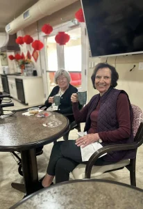 Two older women sit at a table indoors, holding mugs and smiling at the camera, with red decorations hanging in the background, celebrating the Spring Festival.