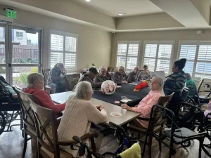 A group of elderly people sit around a table in a well-lit room, engaged in conversation during the Spring Festival; several wheelchairs are visible nearby.