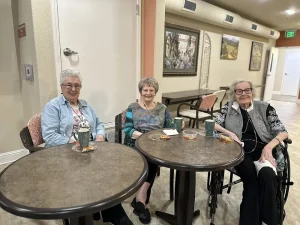 Three elderly women sit at two round tables in a communal room, smiling with drinks and snacks in front of them as they celebrate the Spring Festival together.