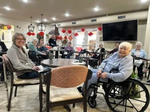 A group of elderly women, many in wheelchairs, sit around tables in a common room decorated with red lanterns for the Lunar New Year, with a large TV on the wall.