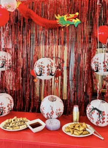 Table with plates of pastries, a bowl of dipping sauce, and a drink set in front of red decorations, paper lanterns with floral designs, and a dragon decoration, perfect for a festive Chinese New Year celebration.