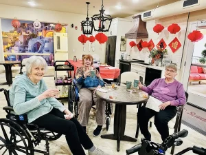 Three elderly women sit around a small table in a decorated room, holding drinks and smiling. Red lanterns and festive decorations hint at the joyful spirit of the Lunar New Year in the background.
