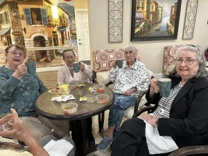 Four elderly women sit around a round table, holding mugs and smiling, enjoying bowls of soup and crackers in a cozy, decorated room as they celebrate the Lunar New Year together.